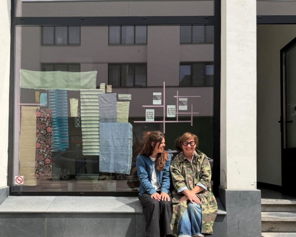 Two women sitting on a step in front of a building with a large window displaying fabric samples. Valerie Tjantele and Katrien Buyle from ATELIER A.PUUR.A 